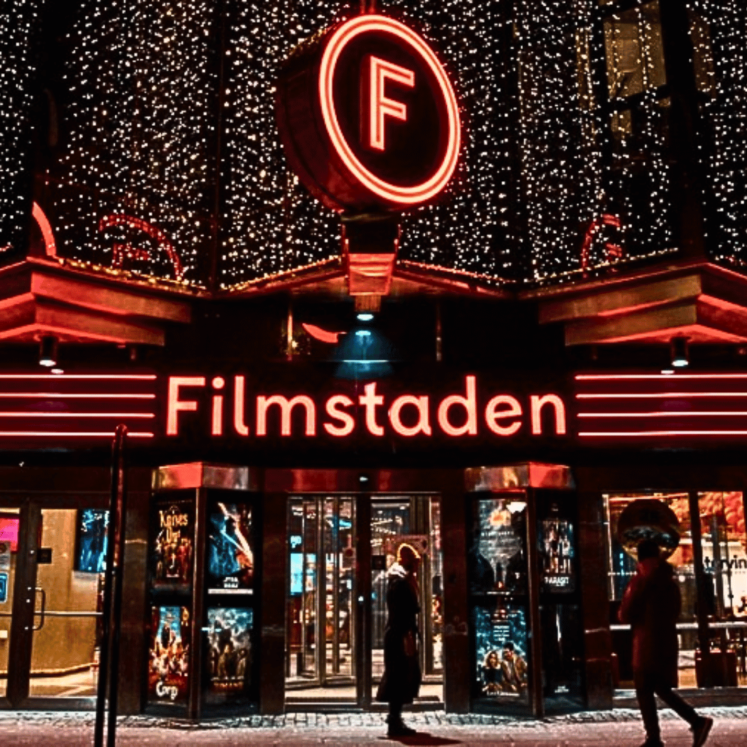 Bild utanför biografen FIlmstadens entré. Två människor står utanför. Det är vinter. Filmstadens entreskylt lyser i röd färg.